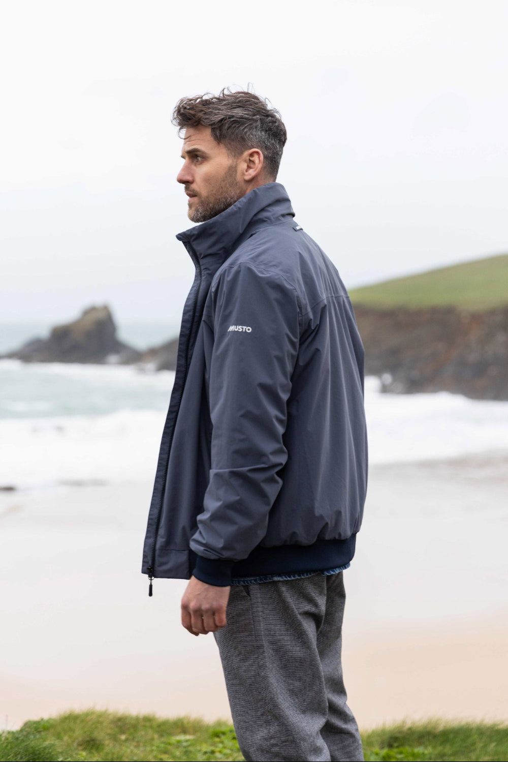 Man wearing a gray jacket with a musto logo standing on a beach.