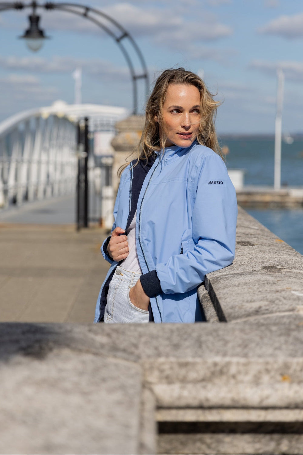 Woman in a blue jacket standing on a dock with a bridge and water in the background